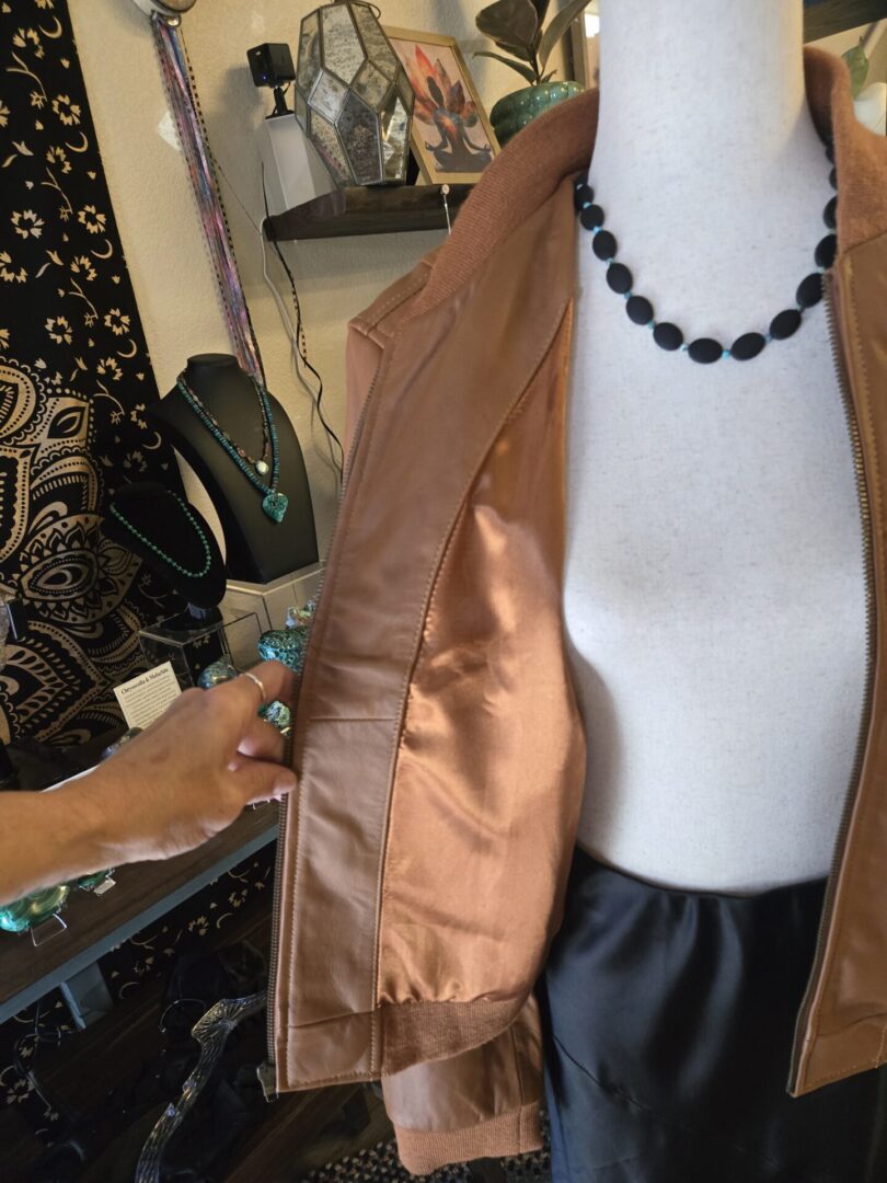 A hand holding open a brown jacket on a mannequin wearing a black necklace and skirt.