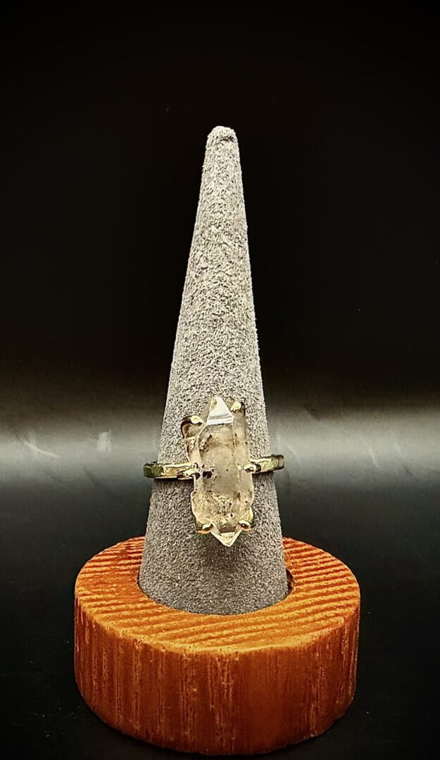A gold ring with a clear, elongated gemstone on a gray cone stand with a wooden base.